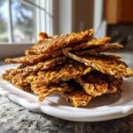 A stack of shiny, golden brown pieces of homemade seed brittle piled high on a white plate.