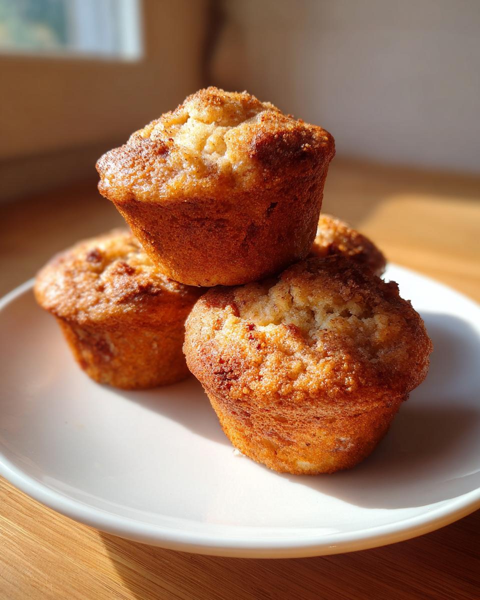 Three golden-brown greek yogurt apple muffins stacked on a white plate, catching warm sunlight.