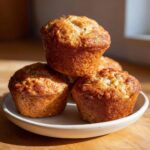 A stack of four golden brown greek yogurt apple muffins resting on a small, light-colored ceramic plate.