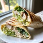 Two halves of a grilled chicken wraps filled with sliced chicken, crisp lettuce, and creamy dressing, resting on a white plate.