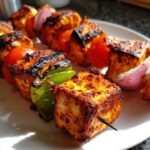 Close-up of beautifully charred paneer and vegetable skewers, similar to chicken kebabs, on a white plate.