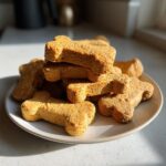 A stack of golden, bone-shaped dog cookies piled high on a light plate, ready for serving.