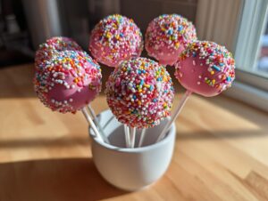 A group of homemade pink starbucks cake pop treats covered in rainbow sprinkles, displayed upright in a white mug.