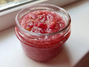 Close-up of a glass jar filled with bright red, chunky rhubarb cinnamon jam sitting on a white surface.