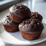 A close-up stack of four rich, dark chocolate muffins topped with mini chocolate chips on a white plate.