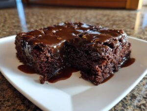 A rich, dark slice of Dr Pepper cake generously topped with a glossy chocolate glaze, served on a white plate.