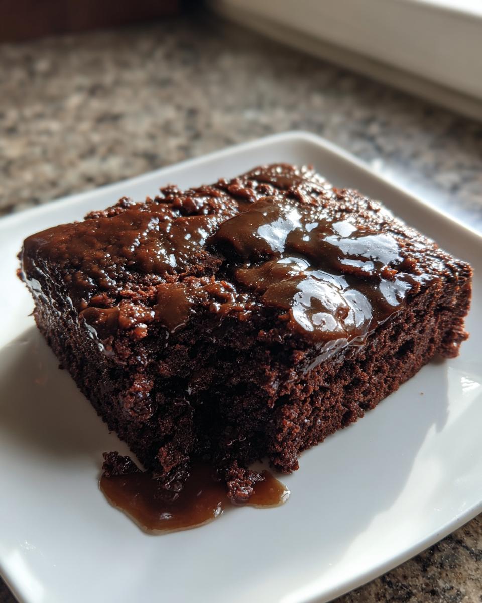 A square slice of rich, dark chocolate Dr Pepper cake topped with a shiny, thick glaze, served on a white plate.