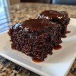 Two squares of moist Dr Pepper cake topped with a thick, glossy chocolate glaze, served on a white plate.