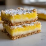 Two stacked squares of vibrant yellow no-bake lemon bars topped with powdered sugar on a white marble surface.