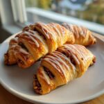 Four golden-brown pecan pie crescents drizzled with white icing, served on a light gray plate indoors.