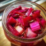 Close-up overhead view of bright pink Pickled Rhubarb with Red Shiso pieces submerged in liquid within a glass jar.