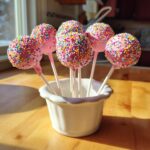 Six pink-coated starbucks cake pop copycat treats covered in rainbow sprinkles standing in a white ceramic holder.