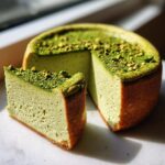 A slice of creamy, pale green pistachio cheesecake next to the main cake, topped with crushed pistachios.