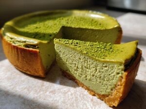 A close-up of a slice of vibrant green pistachio cheesecake next to the main cake, topped with crushed nuts.