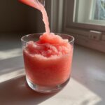 A bright pink frozen strawberry frosé slush being poured into a glass on a sunny windowsill.