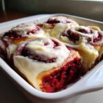 Four freshly baked red velvet cinnamon rolls generously topped with thick white cream cheese frosting in a white baking dish.