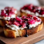A close-up of one piece of Rhubarb and Goat Cheese Bruschetta on a wooden board, topped with creamy cheese and bright pink rhubarb.