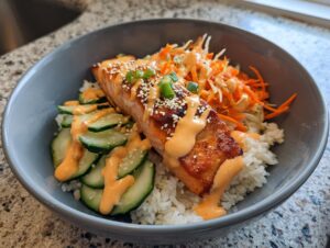 A close-up of a perfectly cooked salmon rice bowl featuring a glazed salmon fillet over white rice, topped with spicy mayo, sesame seeds, sliced cucumbers, and shredded carrots.