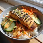 Close-up of a delicious salmon rice bowl featuring a pan-seared salmon fillet over white rice, topped with shredded carrots, cucumber slices, and spicy mayo.