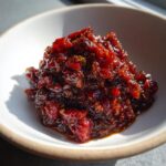 A close-up of rich, dark red, glistening Savory Rhubarb Chutney with Dates piled in a small white bowl.