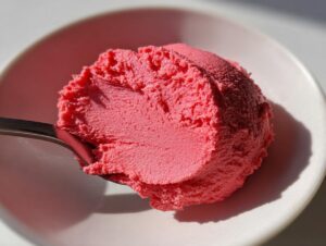 A bright pink scoop of frozen Cinnamon Rhubarb Butter being lifted by a spoon from a white bowl.