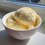 Three scoops of creamy, pale yellow homemade vanilla pudding served in a white bowl, illuminated by sunlight.