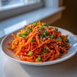 A close-up of a bright orange raw carrot salad, shredded and topped with sesame seeds and chopped green onions.