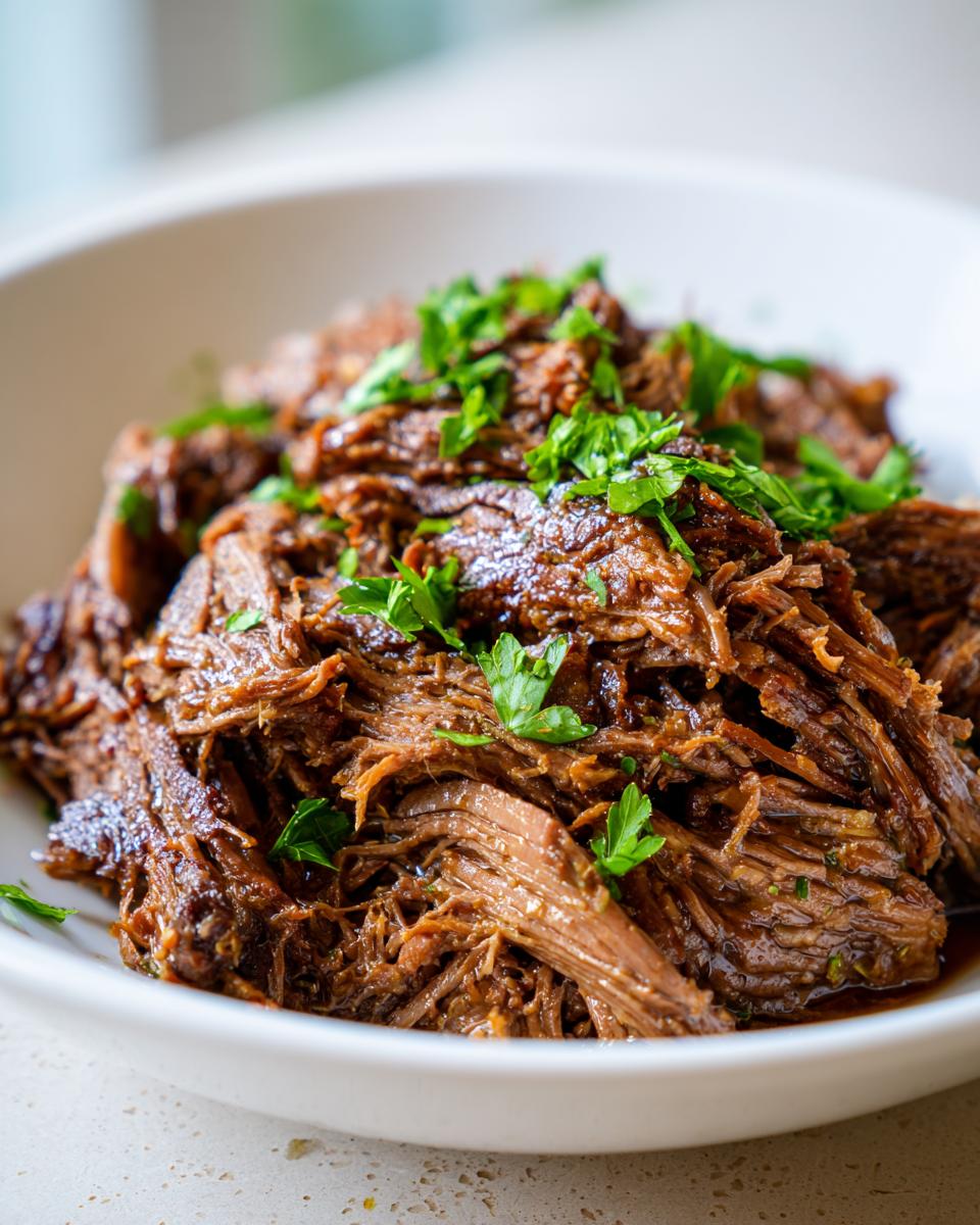 Close-up of tender, shredded slow cooker garlic butter beef garnished with fresh parsley in a white bowl.