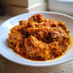 Close-up of tender, shredded meat coated in a rich, orange-red sauce, representing the slow cooker lamb curry.