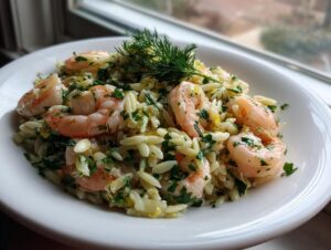 A close-up of a white bowl filled with bright shrimp orzo salad, topped with fresh dill.