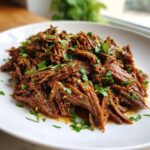 Close-up of tender, shredded slow cooker garlic butter beef, glistening with sauce and topped with fresh parsley.
