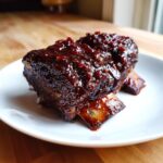 Two pieces of tender Slow-Roasted Rhubarb Short Ribs covered in a thick, dark, glossy glaze, served on a white plate.