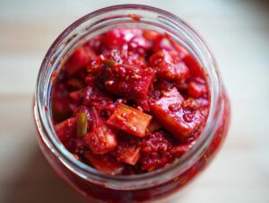 Overhead view of bright red, chopped Small-Batch Rhubarb Kimchi packed tightly in a clear glass jar.