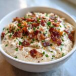 A close-up of a bowl filled with creamy smoked cream cheese dip, generously topped with crumbled bacon and fresh chives.