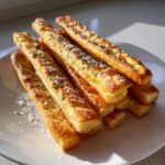 A stack of golden, flaky sourdough discard breadsticks sprinkled with coarse salt and parsley on a white plate.
