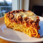 A thick, wedge-shaped slice of spaghetti pie topped with meat sauce and melted cheese.