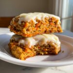 Two stacked carrot cake cookies topped with thick cream cheese frosting, showing shredded carrots inside.