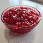 Close-up of bright red Strawberry Rhubarb Pineapple Jam texture in a small clear glass bowl.
