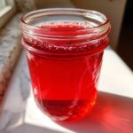 A close-up of vibrant red Strawberry Rhubarb Soda Syrup stored in a clear glass mason jar, catching sunlight.
