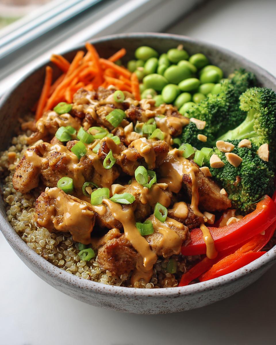A vibrant thai peanut chicken buddha bowl featuring quinoa, chicken drizzled with peanut sauce, broccoli, carrots, edamame, and red peppers.