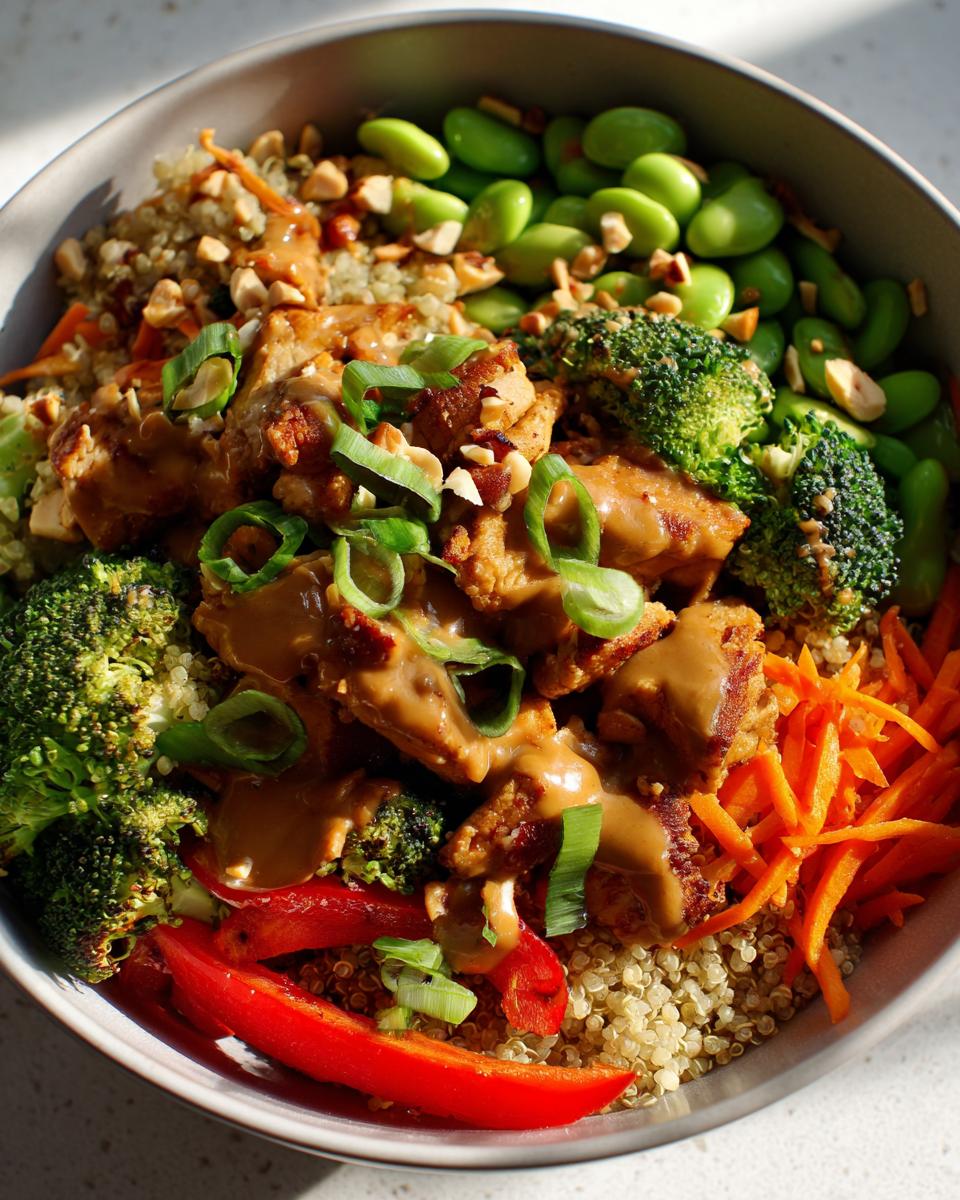 A vibrant thai peanut chicken buddha bowl featuring quinoa, chicken pieces drizzled with peanut sauce, broccoli, edamame, and shredded carrots.