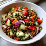 Close-up of a vibrant lentil salad in a white bowl featuring brown lentils, chunks of cucumber, red onion, and bell peppers.