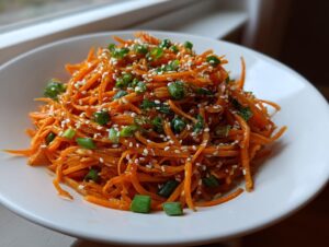 Close-up of a vibrant raw carrot salad made with julienned carrots, topped with white sesame seeds and chopped green onions.