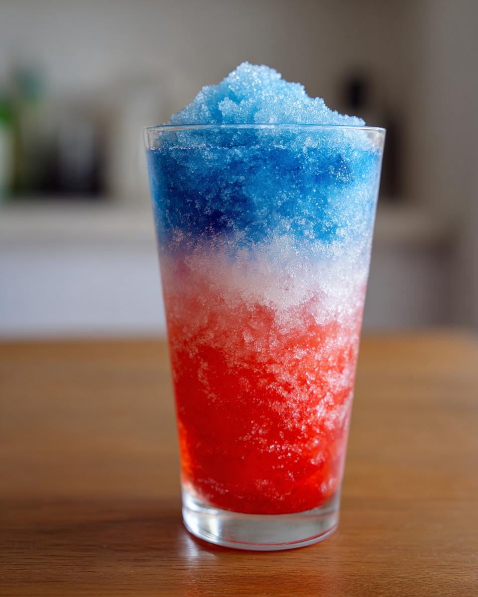 A tall glass filled with a vibrant, layered boozy bomb pop slushie showing red, white, and blue crushed ice.