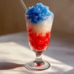 A refreshing boozy bomb pop slushie layered in red, white, and blue crushed ice in a stemmed glass.