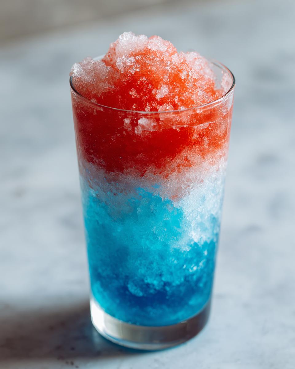 A tall glass filled with a layered, icy boozy bomb pop slushie featuring bright red on top and vibrant blue on the bottom.