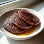 Three dark chocolate oat pancakes stacked on a white plate, drizzled with golden syrup.