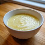 A close-up of a bowl of creamy, pale yellow low calorie potato soup with flecks of green herbs.