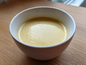 A close-up of a bowl of smooth, creamy yellow low calorie potato soup resting on a light wooden surface.