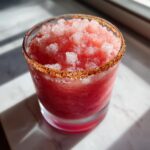 Close-up of a vibrant red frozen firecracker rosé slushie with a chili-sugar rim, sitting on a sunny marble surface.